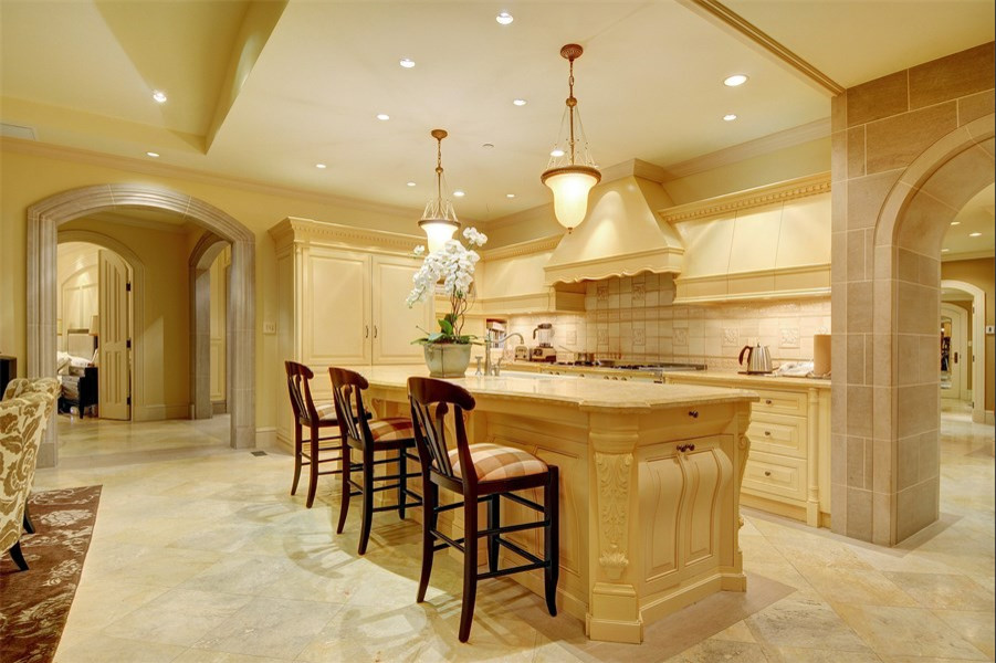 Example of a tuscan limestone floor kitchen design in Vancouver with a farmhouse sink, recessed-panel cabinets, beige cabinets, beige backsplash, stone tile backsplash, paneled appliances and an island