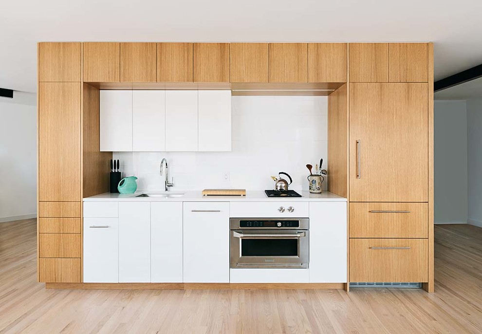 Photo of a small modern single-wall kitchen in New York with a submerged sink, flat-panel cabinets, light wood cabinets, engineered stone countertops, white splashback, ceramic splashback, integrated appliances, light hardwood flooring, no island and white worktops.