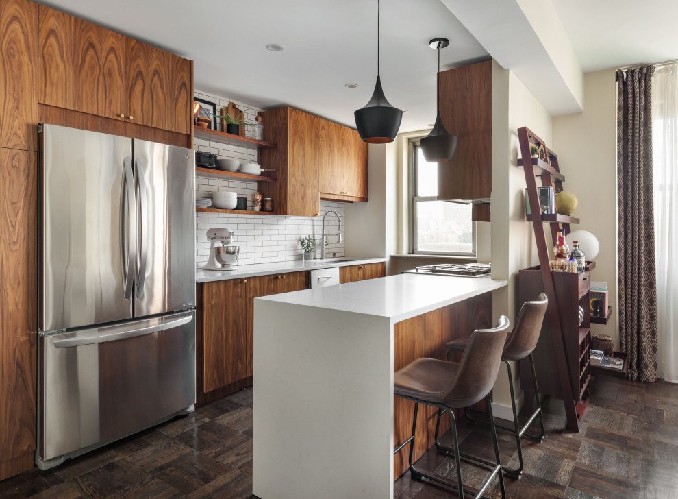 Photo of a contemporary galley kitchen in New York with a submerged sink, flat-panel cabinets, medium wood cabinets, white splashback, stainless steel appliances, dark hardwood flooring, a breakfast bar, brown floors and white worktops.