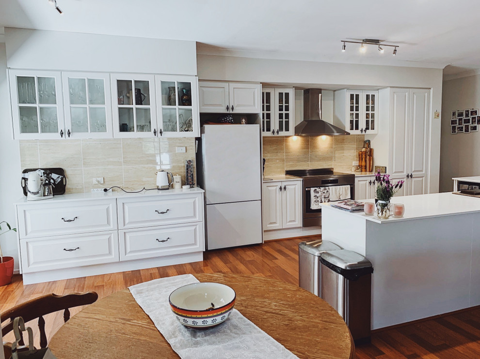 На фото: прямая кухня-гостиная среднего размера в современном стиле с фасадами в стиле шейкер, белыми фасадами, столешницей из акрилового камня, бежевым фартуком, островом и белой столешницей
