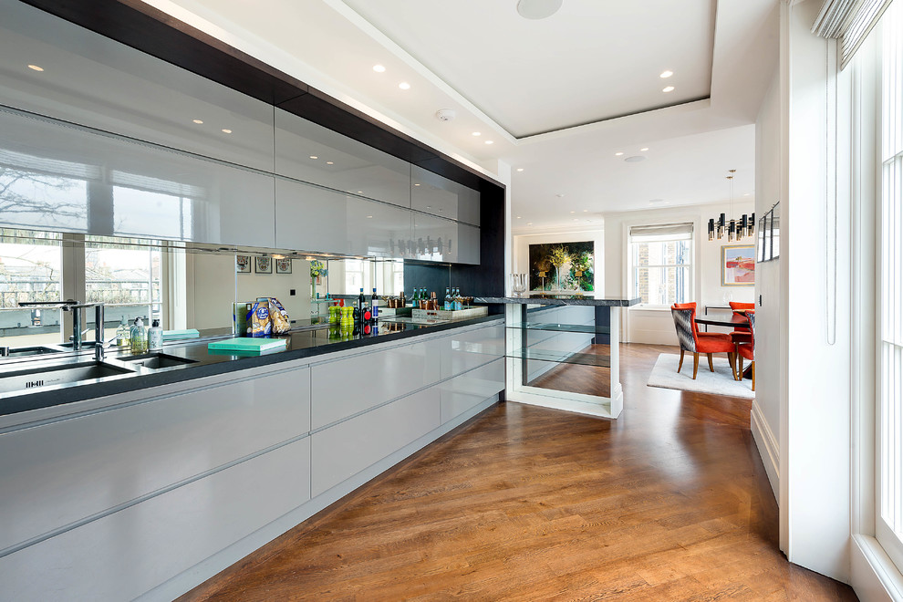Einzeilige Moderne Küche mit Doppelwaschbecken, flächenbündigen Schrankfronten, blauen Schränken, Rückwand aus Spiegelfliesen, braunem Holzboden, braunem Boden und schwarzer Arbeitsplatte in London
