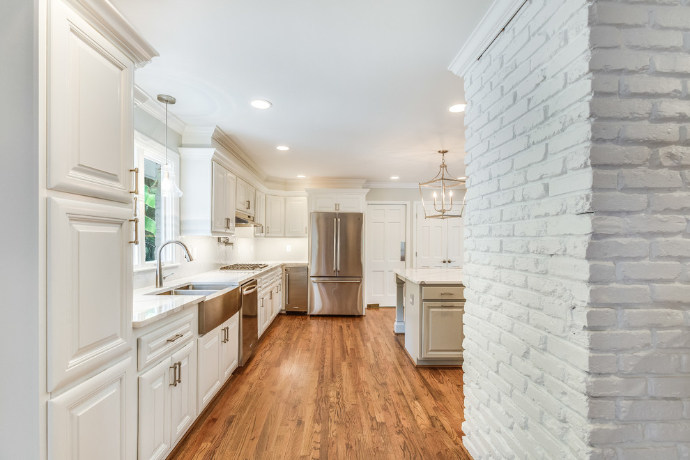 Example of a mid-sized transitional l-shaped medium tone wood floor and brown floor eat-in kitchen design in Birmingham with a farmhouse sink, raised-panel cabinets, white cabinets, quartz countertops, white backsplash, subway tile backsplash, stainless steel appliances, an island and white countertops