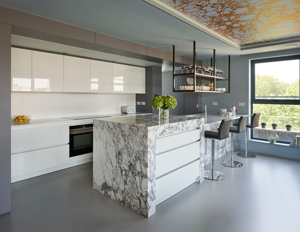 Photo of an expansive contemporary single-wall open plan kitchen in London with flat-panel cabinets, white cabinets, marble worktops, white splashback, stone slab splashback, black appliances, an island, grey floors and white worktops.