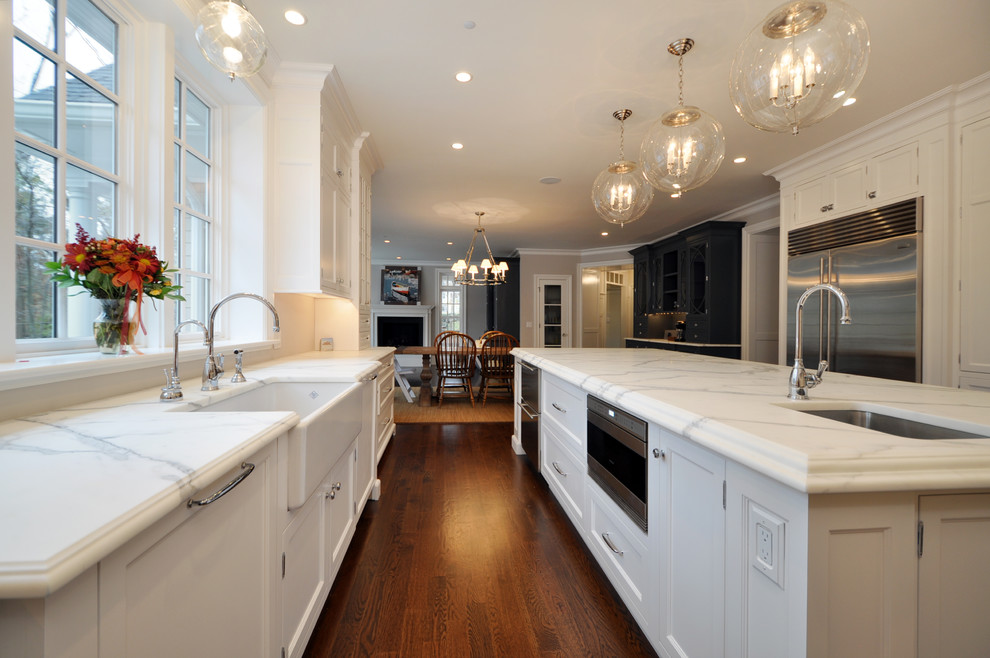 Inredning av ett klassiskt mycket stort vit vitt kök, med en rustik diskho, skåp i shakerstil, grå skåp, marmorbänkskiva, vitt stänkskydd, rostfria vitvaror, mörkt trägolv, en köksö och brunt golv