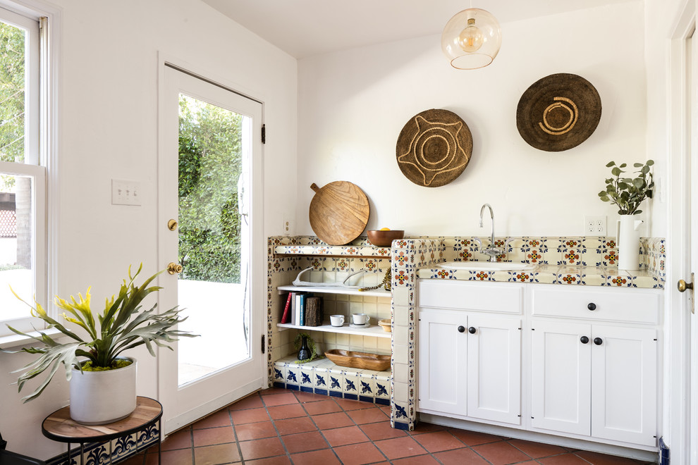 Mediterranean galley kitchen in Los Angeles with a built-in sink, recessed-panel cabinets, white cabinets, tile countertops, beige splashback, terracotta flooring, brown floors and beige worktops.