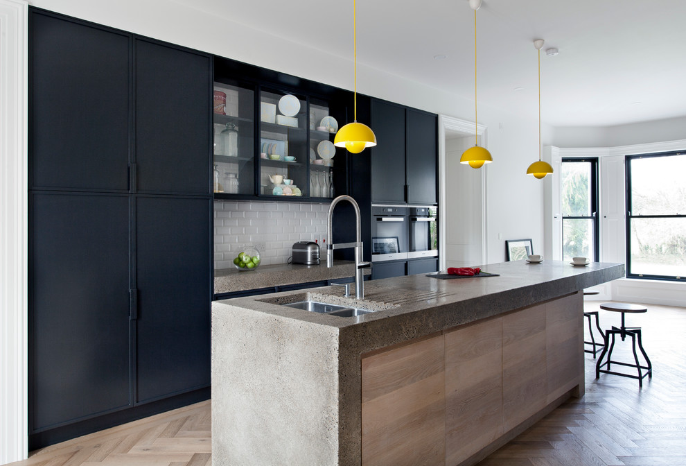 Kitchen - large contemporary light wood floor kitchen idea in Dublin with flat-panel cabinets, concrete countertops, an island, a double-bowl sink, white backsplash, subway tile backsplash and black appliances