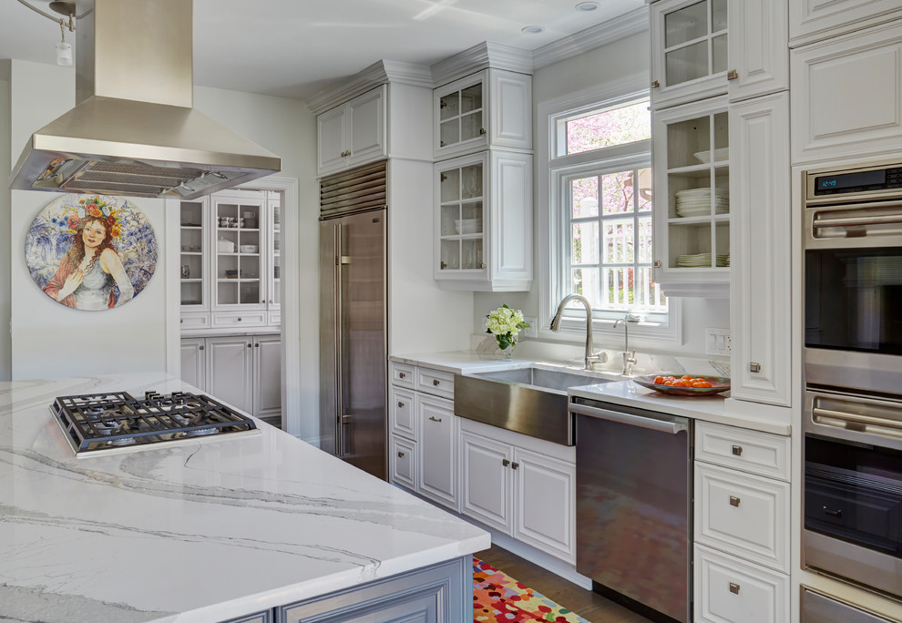 This is an example of a classic galley kitchen in Chicago with a belfast sink, raised-panel cabinets, white cabinets, stainless steel appliances, medium hardwood flooring, an island, brown floors and white worktops.