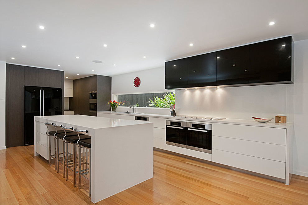 Example of a minimalist galley light wood floor kitchen pantry design in Melbourne with an undermount sink, flat-panel cabinets, white cabinets, quartz countertops, white backsplash, black appliances and an island