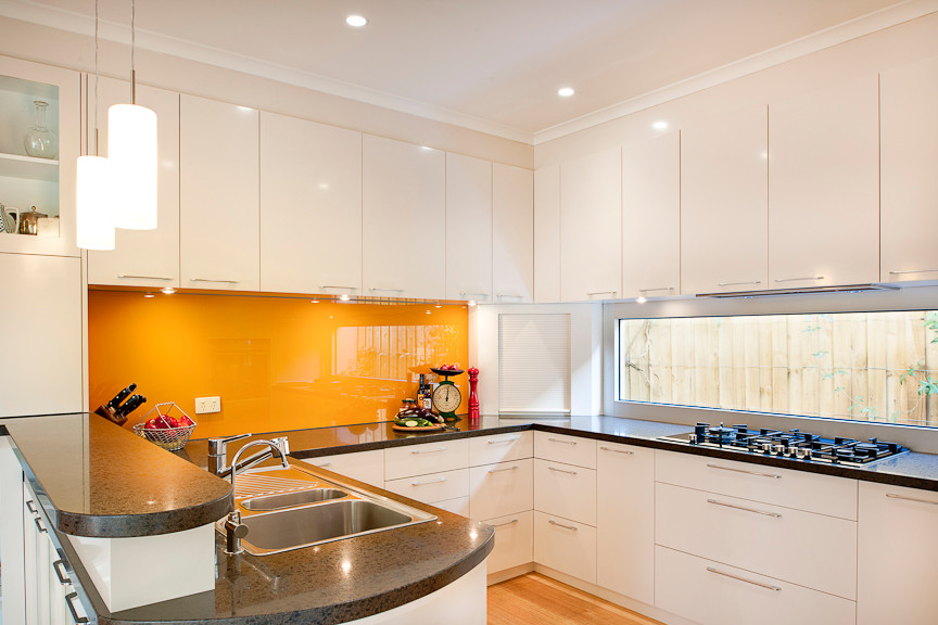 Inredning av ett modernt mellanstort kök, med en halv köksö, släta luckor, vita skåp, bänkskiva i kvarts, orange stänkskydd, glaspanel som stänkskydd, rostfria vitvaror, en nedsänkt diskho och ljust trägolv