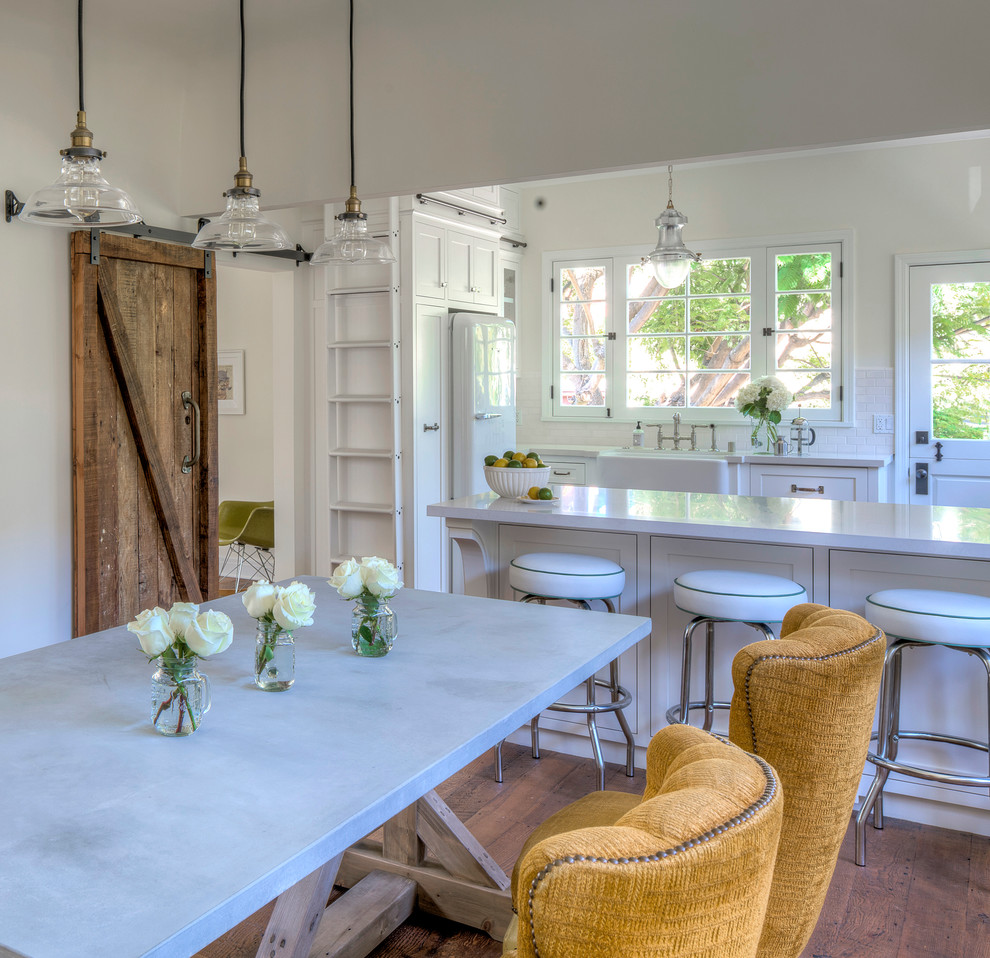 Example of a mid-sized country u-shaped medium tone wood floor eat-in kitchen design in Los Angeles with a farmhouse sink, shaker cabinets, white cabinets, solid surface countertops, white backsplash, subway tile backsplash, stainless steel appliances and a peninsula