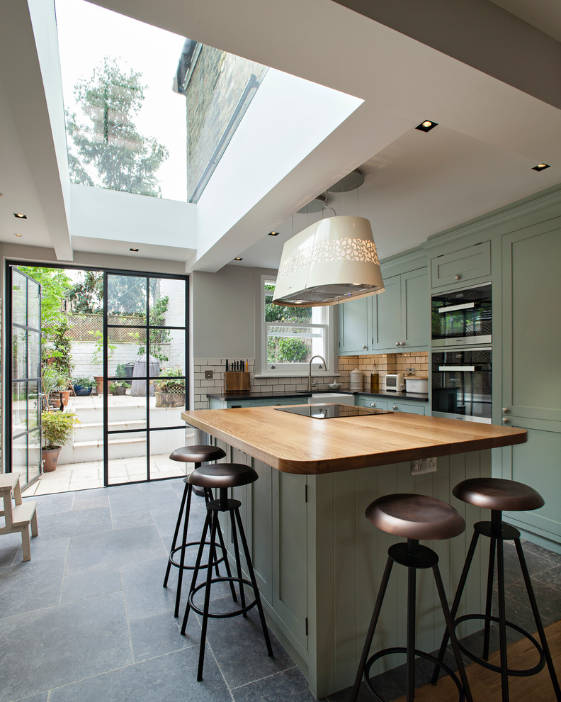 Medium sized contemporary l-shaped kitchen in London with a belfast sink, shaker cabinets, green cabinets, wood worktops, white splashback, metro tiled splashback, black appliances, ceramic flooring and an island.