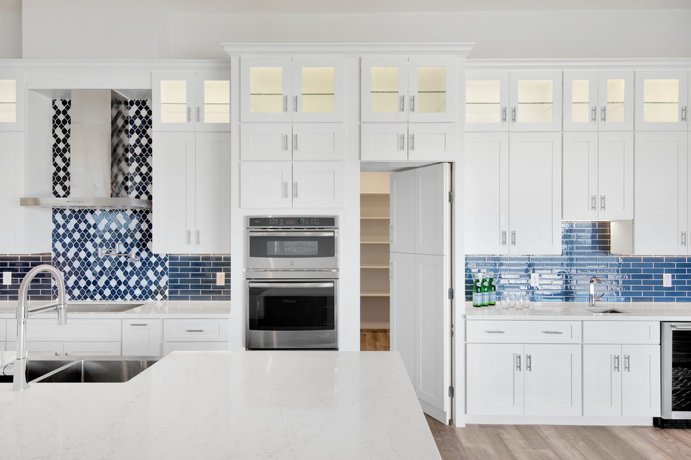 This is an example of a large modern l-shaped kitchen pantry in Phoenix with a submerged sink, shaker cabinets, white cabinets, engineered stone countertops, blue splashback, ceramic splashback, stainless steel appliances, medium hardwood flooring, an island, brown floors and white worktops.