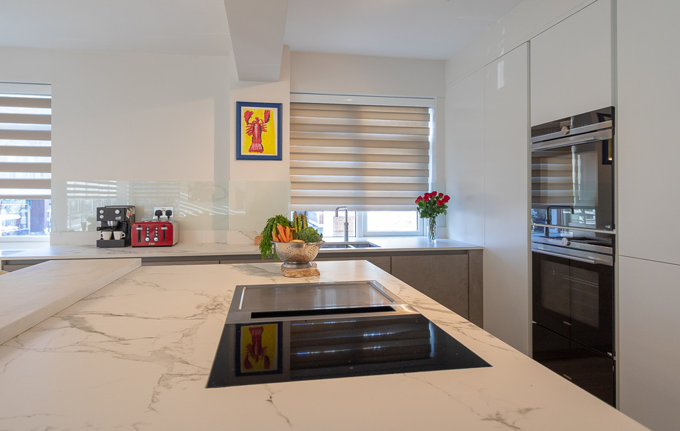 Photo of a medium sized modern l-shaped open plan kitchen in Surrey with an integrated sink, flat-panel cabinets, white cabinets, composite countertops, white splashback, glass sheet splashback, black appliances, medium hardwood flooring, an island, brown floors and white worktops.