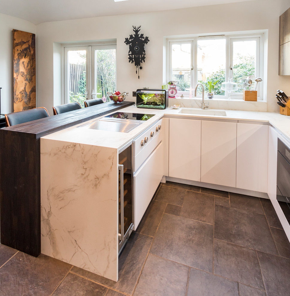 This is an example of a small modern u-shaped open plan kitchen in London with an integrated sink, flat-panel cabinets, white cabinets, composite countertops, metallic splashback, mirror splashback, black appliances, ceramic flooring, a breakfast bar and brown floors.