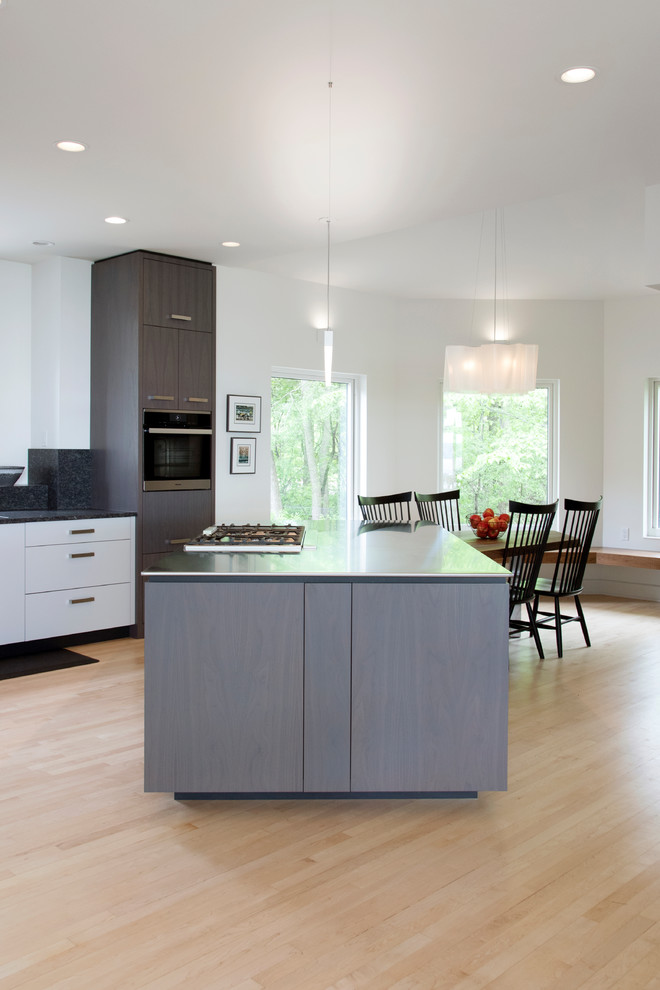 Inspiration for a large modern l-shaped kitchen in Minneapolis with a single-bowl sink, flat-panel cabinets, white cabinets, stainless steel worktops, black splashback, stone slab splashback, integrated appliances, light hardwood flooring and an island.