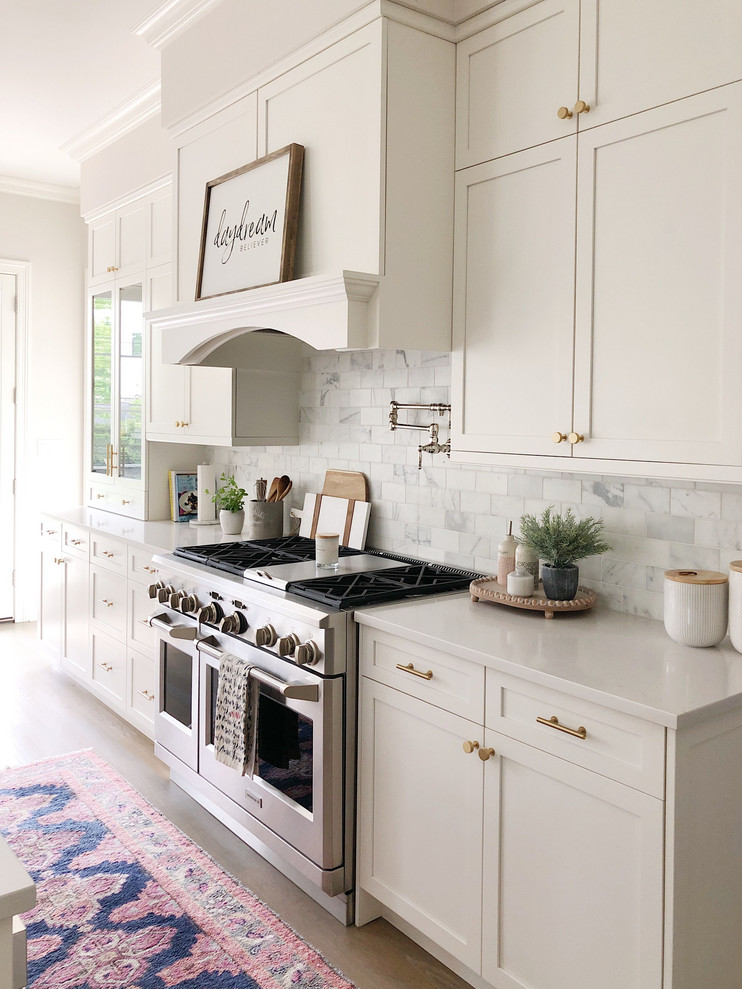 Inspiration for a large classic kitchen in Columbus with a belfast sink, recessed-panel cabinets, white cabinets, engineered stone countertops, white splashback, stone tiled splashback, stainless steel appliances, medium hardwood flooring, an island, brown floors and white worktops.
