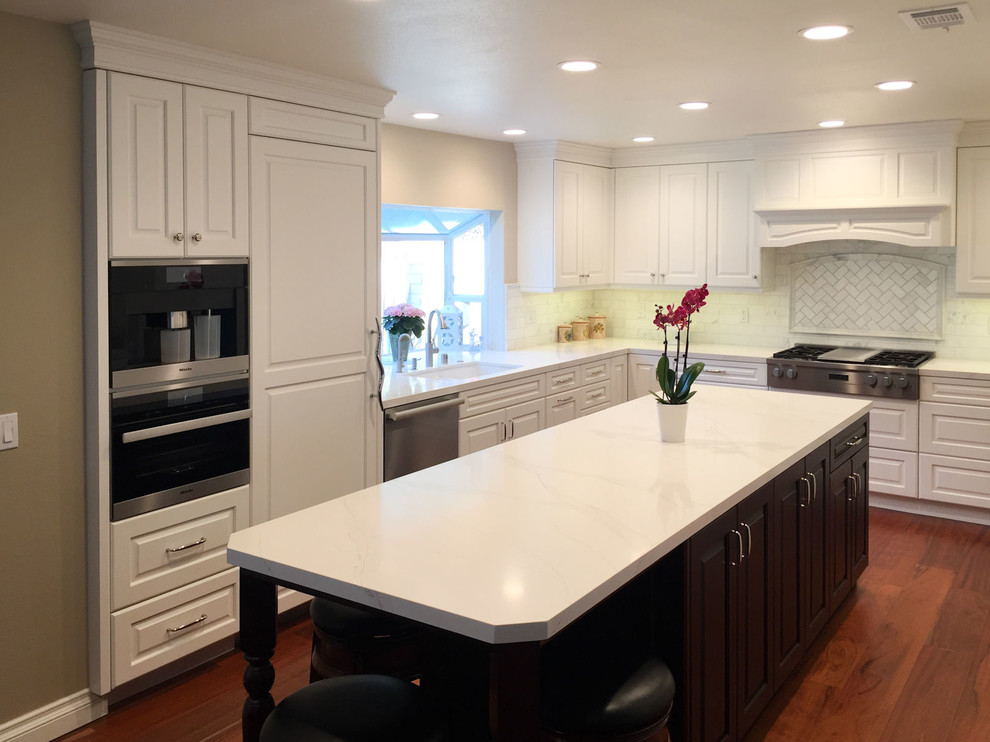 Inspiration for a mid-sized timeless u-shaped medium tone wood floor and brown floor eat-in kitchen remodel in Los Angeles with an undermount sink, raised-panel cabinets, white cabinets, quartz countertops, beige backsplash, stainless steel appliances, an island and white countertops