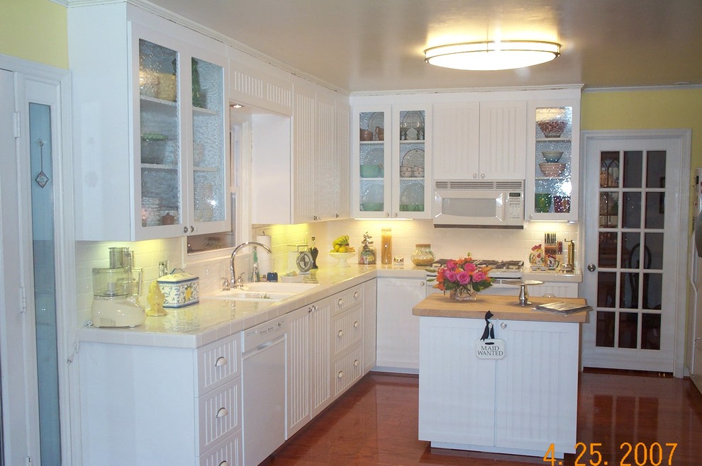 Foto på ett mellanstort lantligt kök, med en dubbel diskho, luckor med glaspanel, vita skåp, kaklad bänkskiva, vitt stänkskydd, stänkskydd i tunnelbanekakel, vita vitvaror, mellanmörkt trägolv och en köksö