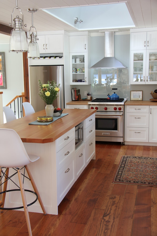 Bild på ett mellanstort lantligt l-kök, med skåp i shakerstil, vita skåp, träbänkskiva, blått stänkskydd, glaspanel som stänkskydd, rostfria vitvaror, mellanmörkt trägolv, en köksö och en rustik diskho