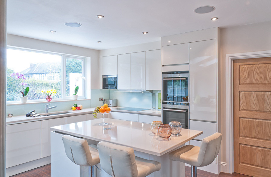 Photo of a small modern l-shaped kitchen/diner in London with a submerged sink, flat-panel cabinets, white cabinets, quartz worktops, white splashback, glass sheet splashback, stainless steel appliances, medium hardwood flooring and an island.