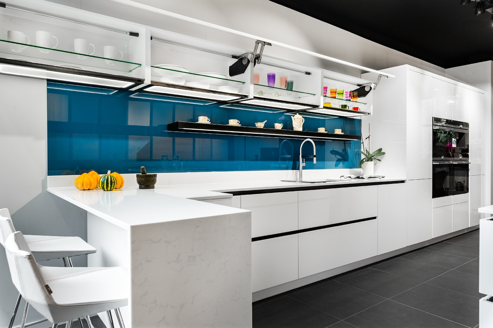 Design ideas for a large modern single-wall open plan kitchen in London with a built-in sink, glass-front cabinets, white cabinets, quartz worktops, blue splashback, glass sheet splashback, integrated appliances, porcelain flooring, a breakfast bar, grey floors and white worktops.