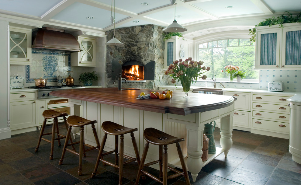 Klassisk inredning av ett kök, med en rustik diskho, luckor med glaspanel, beige skåp, vitt stänkskydd, rostfria vitvaror och skiffergolv