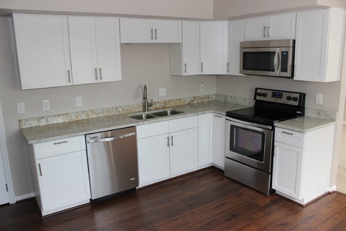 Compact L-shaped kitchen with white shaker RTA cabinets, light granite countertops and short backsplash, stainless stove, microwave and dishwasher, and dark wood floors.