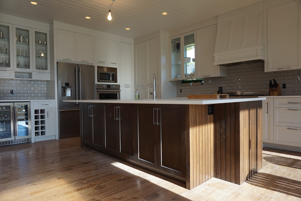 Photo of a medium sized classic l-shaped kitchen in Calgary with grey splashback, metro tiled splashback, a submerged sink, shaker cabinets, white cabinets, engineered stone countertops, stainless steel appliances, medium hardwood flooring, an island and brown floors.