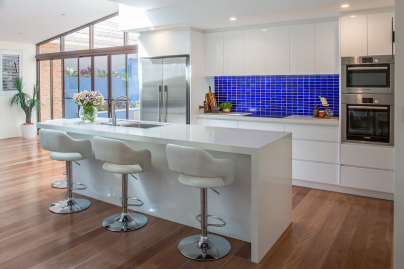 This is an example of a medium sized contemporary galley kitchen/diner in Other with a submerged sink, flat-panel cabinets, white cabinets, engineered stone countertops, blue splashback, glass tiled splashback, stainless steel appliances, medium hardwood flooring and an island.