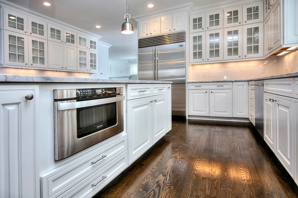 Mid-sized transitional l-shaped dark wood floor open concept kitchen photo in New York with an undermount sink, raised-panel cabinets, white cabinets, marble countertops, white backsplash, porcelain backsplash, stainless steel appliances and an island
