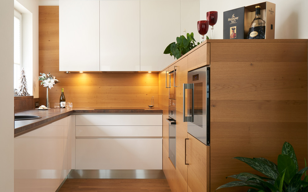 Bild på ett avskilt, litet funkis l-kök, med en nedsänkt diskho, släta luckor, vita skåp, laminatbänkskiva, brunt stänkskydd, rostfria vitvaror och ljust trägolv