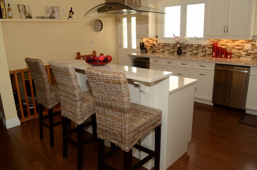Bild på ett kök, med en undermonterad diskho, luckor med infälld panel, vita skåp, bänkskiva i kvartsit, grått stänkskydd, stänkskydd i glaskakel, mörkt trägolv och en köksö