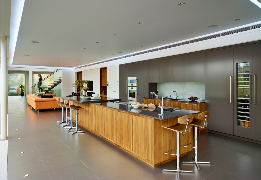 На фото: параллельная кухня-гостиная в стиле модернизм с врезной мойкой, плоскими фасадами, серыми фасадами, фартуком из стекла, техникой под мебельный фасад и островом