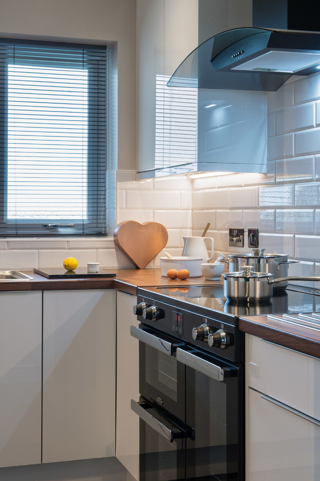 Photo of a medium sized modern u-shaped enclosed kitchen in Cornwall with an integrated sink, glass-front cabinets, white cabinets, wood worktops, white splashback, cement tile splashback, limestone flooring, an island, black floors and brown worktops.