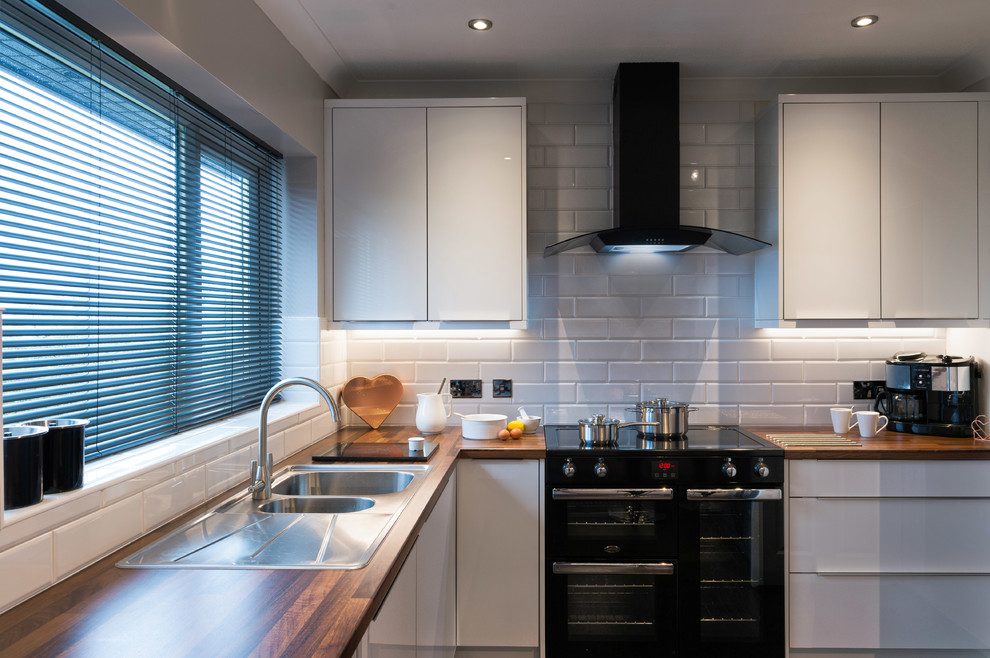 Design ideas for a medium sized modern u-shaped enclosed kitchen in Cornwall with an integrated sink, glass-front cabinets, white cabinets, wood worktops, white splashback, cement tile splashback, limestone flooring, an island, black floors and brown worktops.