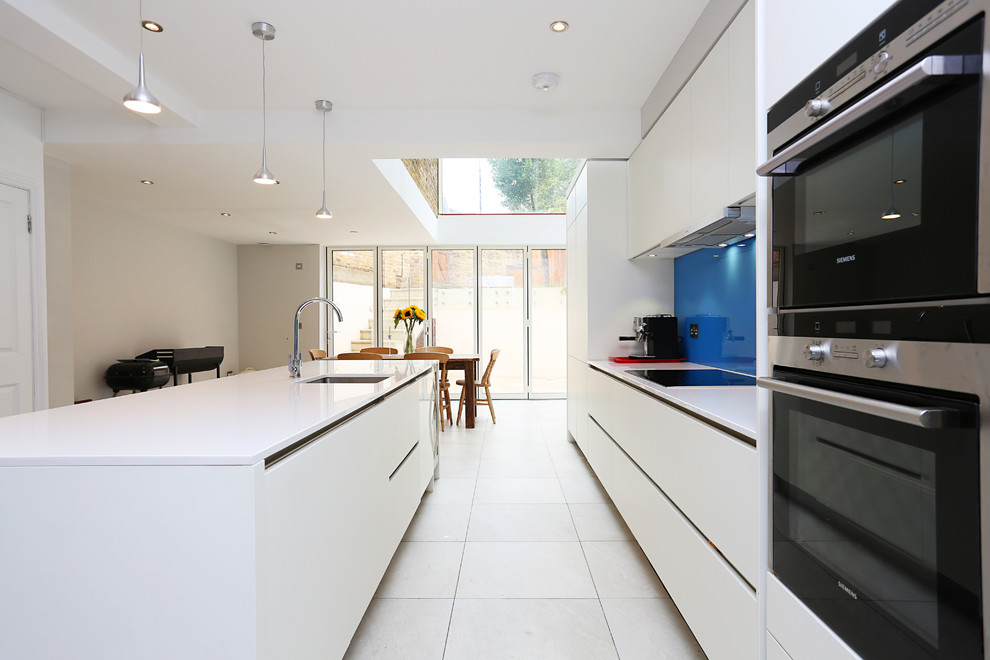На фото: кухня-гостиная в стиле модернизм с плоскими фасадами, белыми фасадами, синим фартуком, фартуком из стекла, техникой из нержавеющей стали, полом из керамогранита и островом с
