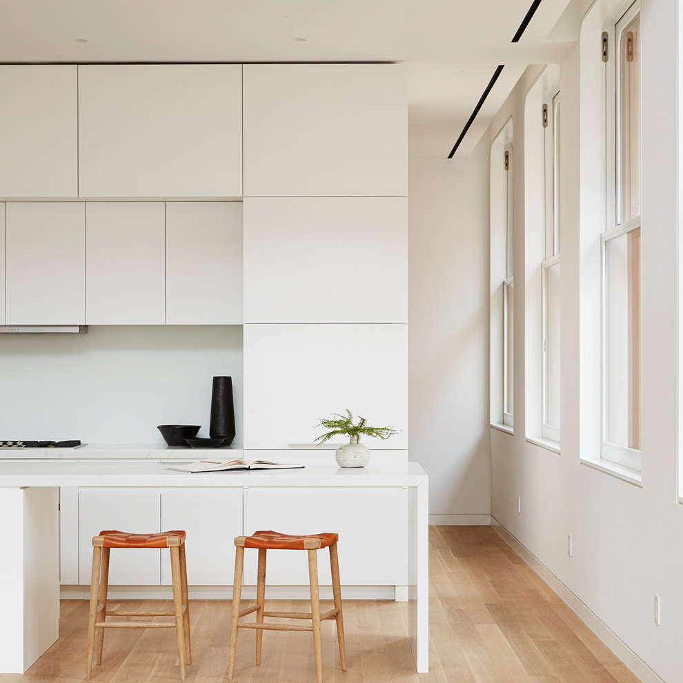 Réalisation d'une cuisine parallèle minimaliste avec parquet clair, un placard à porte plane, des portes de placard blanches, îlot, un sol beige et un plan de travail blanc.