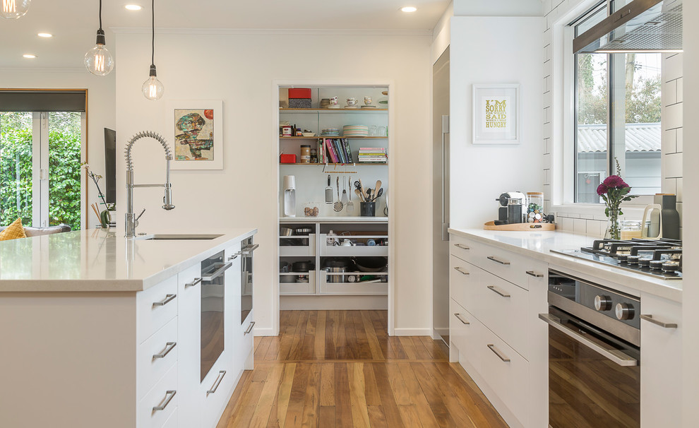 Example of a mid-sized trendy single-wall light wood floor and beige floor open concept kitchen design in Auckland with an undermount sink, flat-panel cabinets, white cabinets, quartz countertops, white backsplash, subway tile backsplash, stainless steel appliances, an island and white countertops