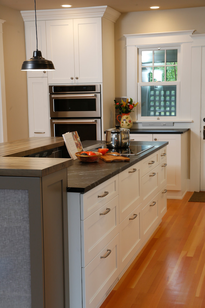 Mid-sized transitional l-shaped medium tone wood floor eat-in kitchen photo in Seattle with a farmhouse sink, shaker cabinets, white cabinets, soapstone countertops, brown backsplash, ceramic backsplash, stainless steel appliances and an island