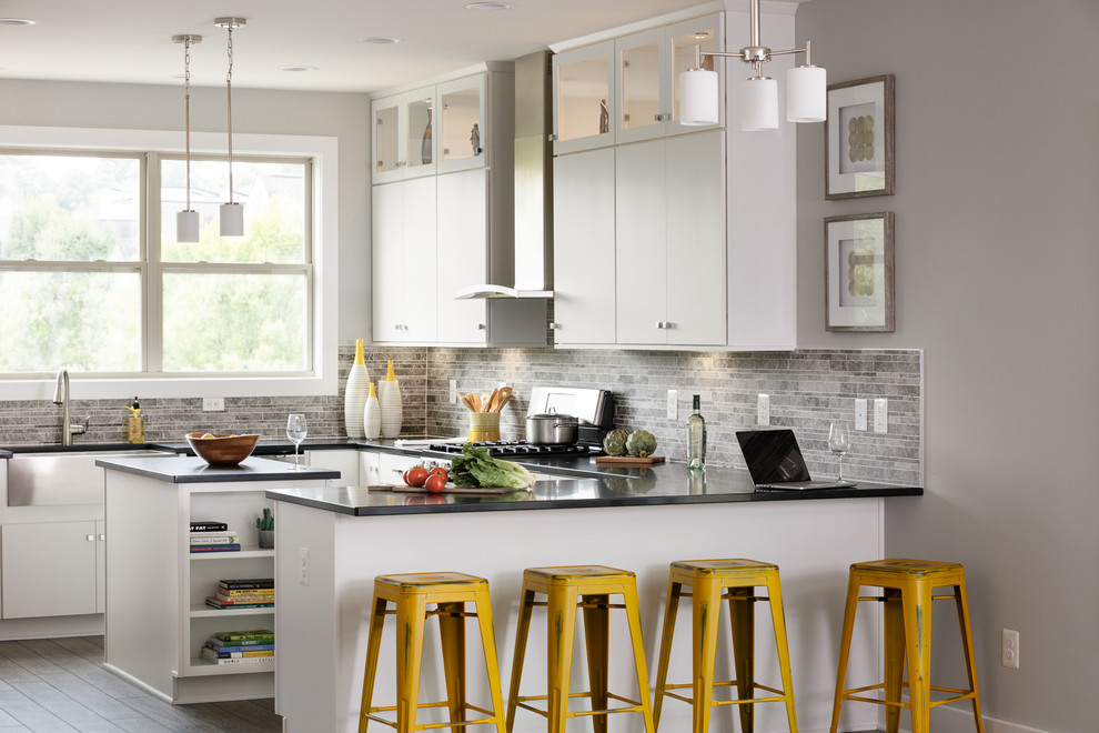 Example of a mid-sized trendy u-shaped laminate floor and brown floor open concept kitchen design in Other with a farmhouse sink, flat-panel cabinets, white cabinets, quartz countertops, gray backsplash, stone tile backsplash, stainless steel appliances, an island and gray countertops