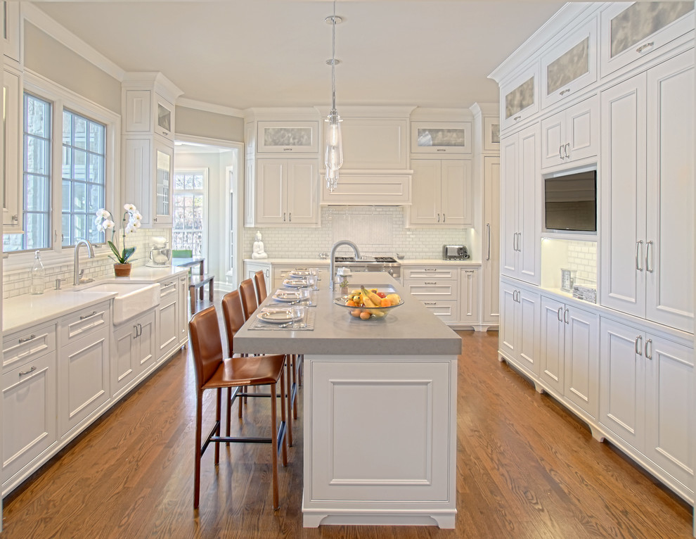Klassisk inredning av ett mellanstort vit vitt kök, med en rustik diskho, luckor med infälld panel, vita skåp, bänkskiva i kvarts, vitt stänkskydd, stänkskydd i porslinskakel, rostfria vitvaror, mellanmörkt trägolv, en köksö och brunt golv