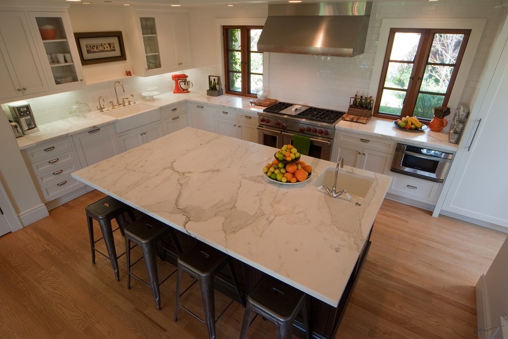 Example of a large transitional l-shaped medium tone wood floor open concept kitchen design in Orange County with a farmhouse sink, shaker cabinets, white cabinets, marble countertops, white backsplash, ceramic backsplash, stainless steel appliances and an island
