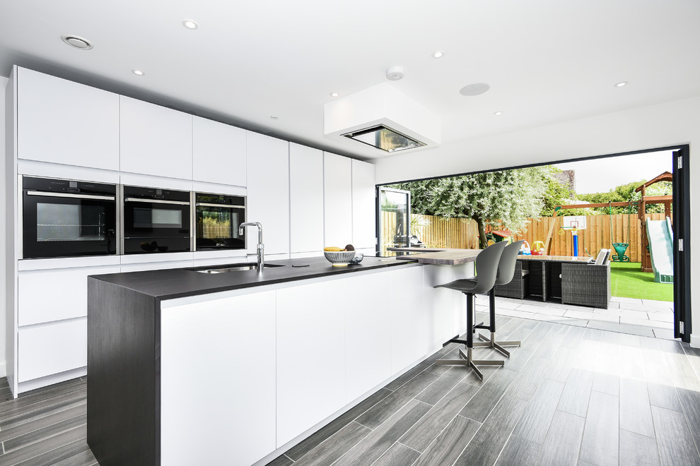 Design ideas for a medium sized contemporary single-wall kitchen in Buckinghamshire with a double-bowl sink, flat-panel cabinets, white cabinets, stainless steel appliances, an island, grey floors and black worktops.