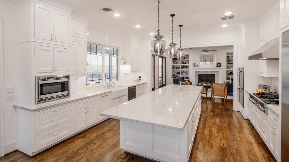 Huge transitional u-shaped medium tone wood floor and brown floor enclosed kitchen photo in Houston with a farmhouse sink, recessed-panel cabinets, white cabinets, quartz countertops, white backsplash, stainless steel appliances, an island and white countertops