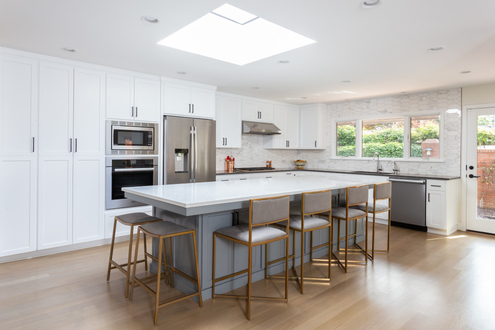 Galley kitchen/diner in Portland with a built-in sink, recessed-panel cabinets, white cabinets, marble worktops, white splashback, ceramic splashback, stainless steel appliances, light hardwood flooring, an island, brown floors and white worktops.