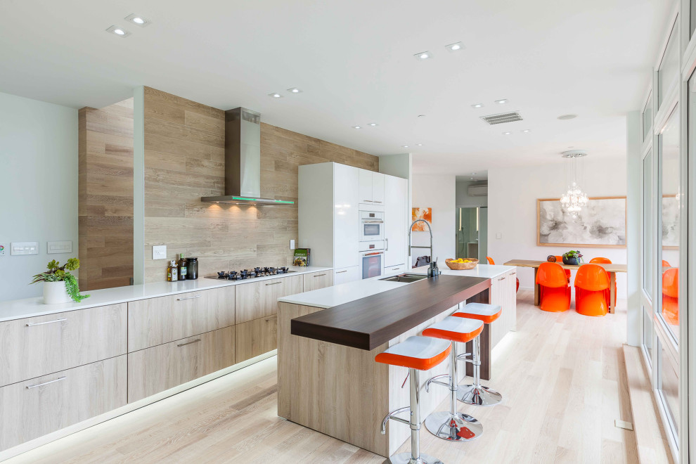 Contemporary galley kitchen in Seattle with a submerged sink, flat-panel cabinets, light wood cabinets, beige splashback, white appliances, light hardwood flooring, an island, beige floors and white worktops.