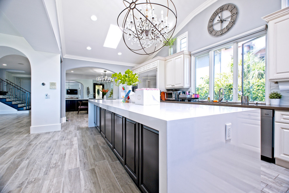 Large modern l-shaped open plan kitchen in Orange County with a built-in sink, raised-panel cabinets, engineered stone countertops, white splashback, porcelain splashback, stainless steel appliances, porcelain flooring, an island, beige floors and white worktops.