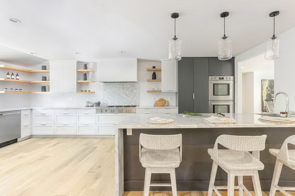 Modern inredning av ett stort vit vitt kök, med en undermonterad diskho, släta luckor, vita skåp, bänkskiva i kvartsit, grått stänkskydd, stänkskydd i sten, rostfria vitvaror, ljust trägolv, en halv köksö och beiget golv