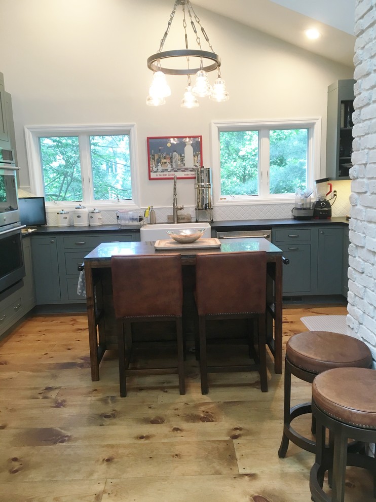 This is an example of a small country u-shaped kitchen/diner in New York with a belfast sink, blue cabinets, white splashback, ceramic splashback, stainless steel appliances, light hardwood flooring and an island.