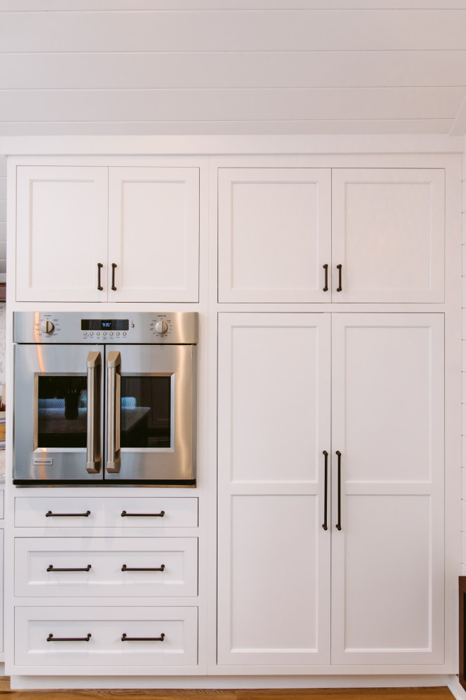 Bild på ett stort maritimt vit vitt kök, med en rustik diskho, luckor med profilerade fronter, vita skåp, bänkskiva i kvarts, vitt stänkskydd, rostfria vitvaror, mellanmörkt trägolv, en köksö och brunt golv